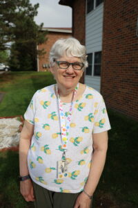 Denise poses for a photo standing outside. She is wearing a floral t-shirt and has white hair and glasses.