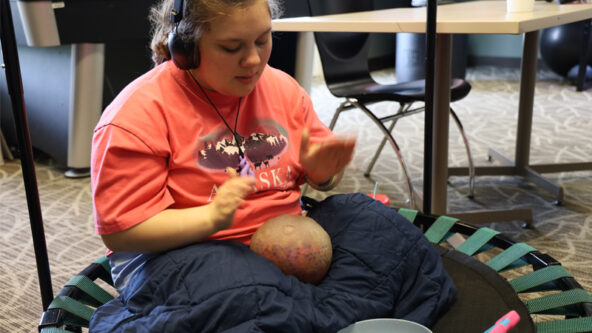 Savannah wears an orange t-shirt and headphones. She sits on a mini trampoline surrounded by a squishy ball, blankets, snacks, and an ipad.