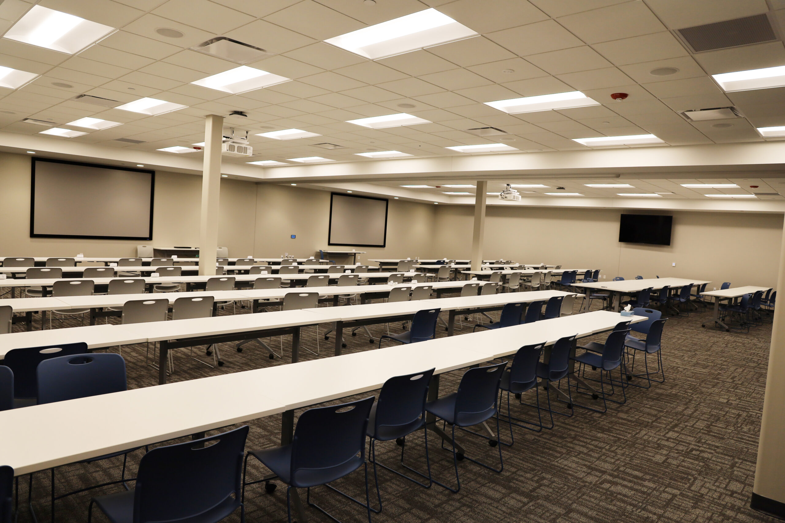 A large white and grey open space with TV screens on the wall and 40 tables and chairs lined up.