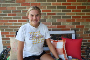 Abby sits on her front porch wearing a white t-shirt and shirts. Her blonde hair is pulled into a ponytail.