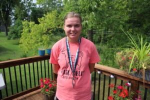 Sarah poses for a photo standing on her back deck. She is wearing a blue lanyard, coral t-shirt with a dog, and has light blondish-brown hair pulled into a ponytail.