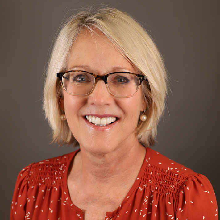 A light skin woman with cropped blonde hair, blue eyes, and glasses. She is wearing a red shirt with pleats at the shoulder.