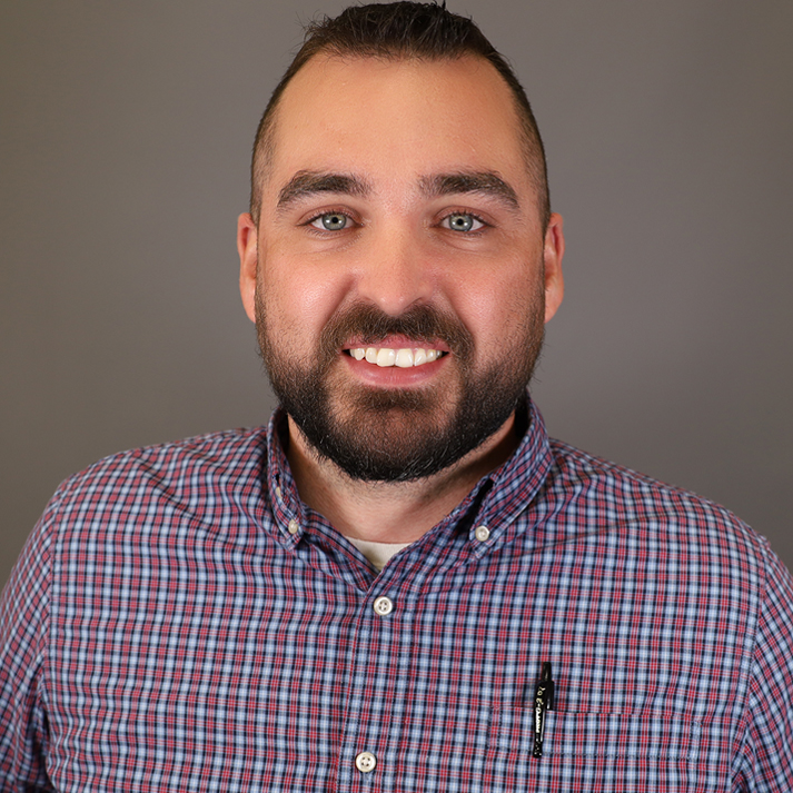 A man with brown hair and trimmed beard. He has blue eyes, and is wearing a plaid button up.